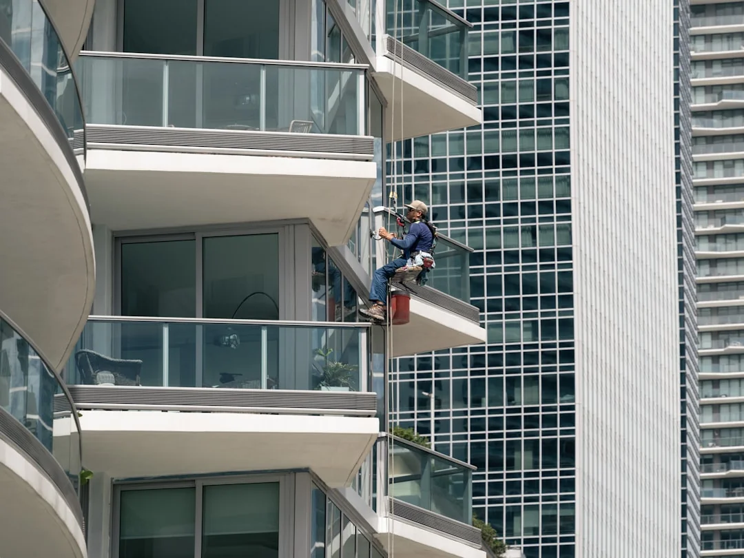 services-02 Black and white from below of man steeplejack in protective helmet with professional equipment cleaning glass wall of modern tower